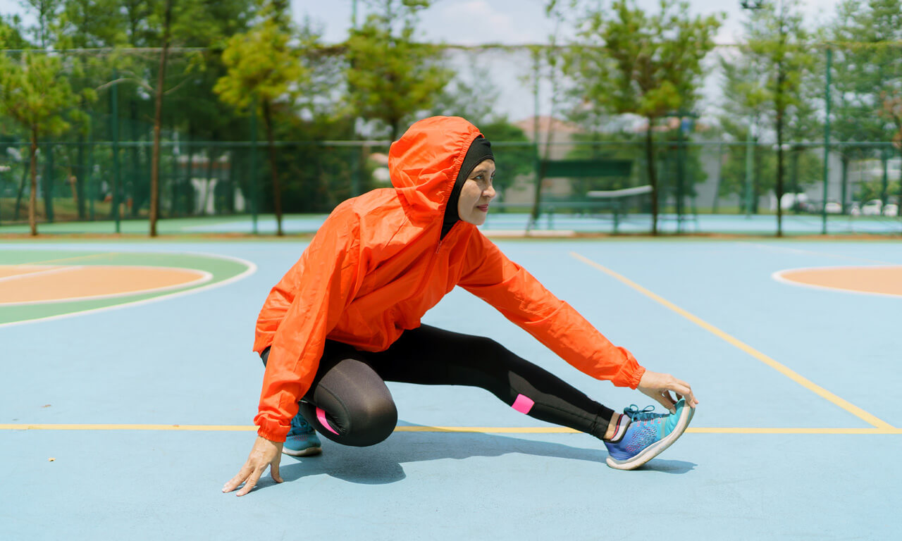A women wearing an orange jacket, on a running track, kneeling on one knee and streching her other knee