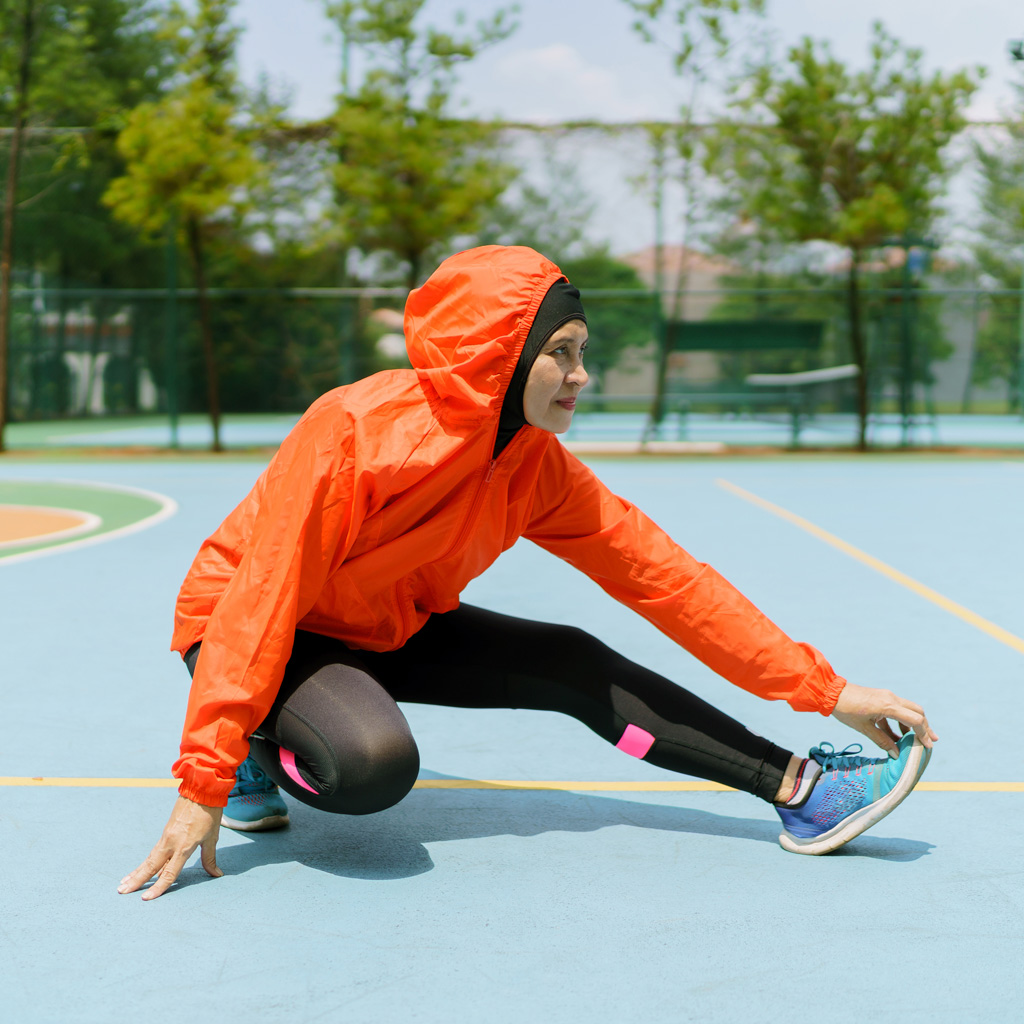 A women wearing an orange jacket, on a running track, kneeling on one knee and streching her other knee A women wearing an orange jacket, on a running track, kneeling on one knee and streching her other knee