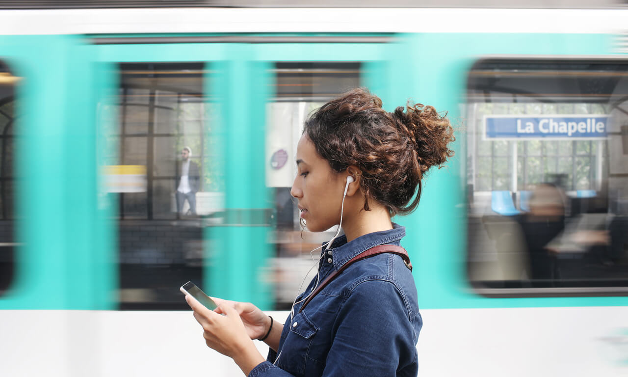 Women with brown hair in a bun, wearing a denim jacket, walking past a blue shop, looking at her phone, wearing wired earphones