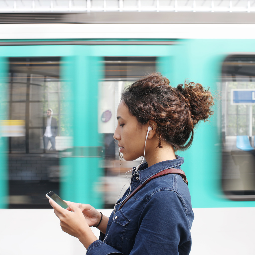 Women with brown hair in a bun, wearing a denim jacket, walking past a blue shop, looking at her phone, wearing wired earphones Women with brown hair in a bun, wearing a denim jacket, walking past a blue shop, looking at her phone, wearing wired earphones