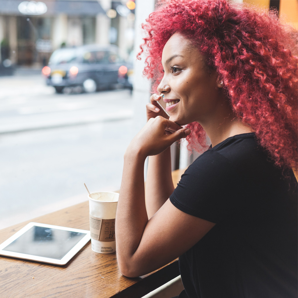 A lady wearing a black t-shirt, holding her phone to her ear and smiling, sitting in a coffee shop window, with a coffee and an IPad in front of her A lady wearing a black t-shirt, holding her phone to her ear and smiling, sitting in a coffee shop window, with a coffee and an IPad in front of her