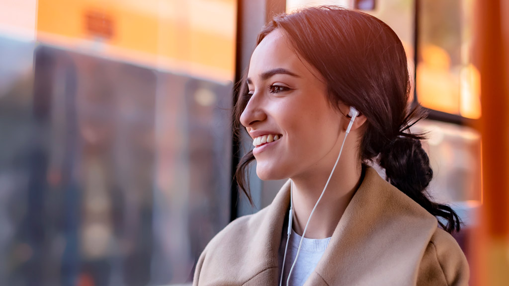 A lady wearing a beige coat, sitting on public transport, wearing wired earphones, and looking out the window smiling