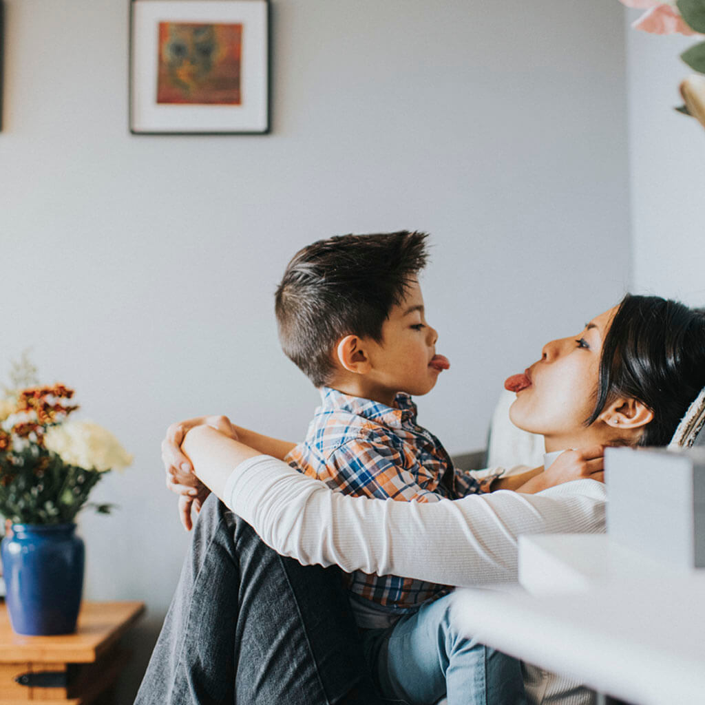 mum and son, sitting on her lap on a white sofa, sticking their tongues out at each other