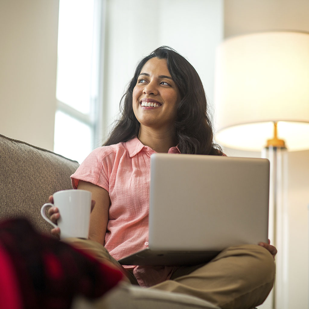 Woman, with brown hair, wearing a pink shirt, sitting on a brown sofa, holding a white mug and silver laptop, looking out of a window