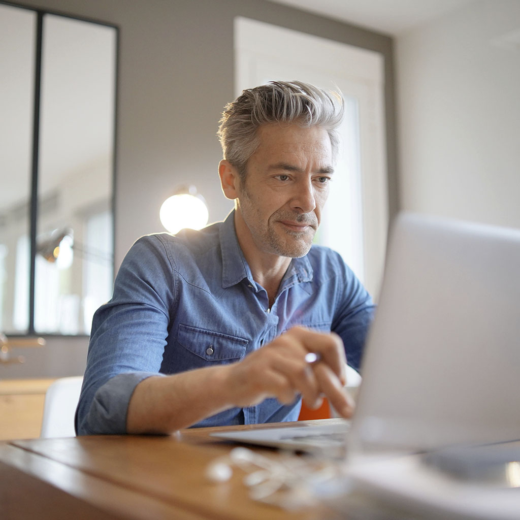 man with silver/grey hair, wearing a blue shirt, sitting at a wooden desk with a light a square mirror behind him, about to type of his silver laptop.