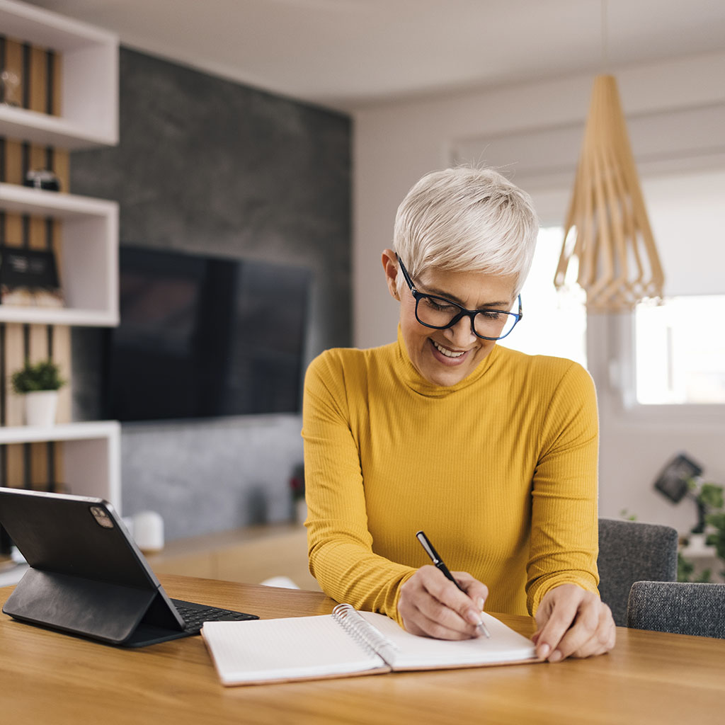 young woman, with short platinum blonde hair, wearing a bright yellow polo next and black rimmed glasses, sits at a wooden table writing in her notepad. A black iPad is to her right and there is a TV on a grey wall in the background. young woman, with short platinum blonde hair, wearing a bright yellow polo next and black rimmed glasses, sits at a wooden table writing in her notepad. A black iPad is to her right and there is a TV on a grey wall in the background.