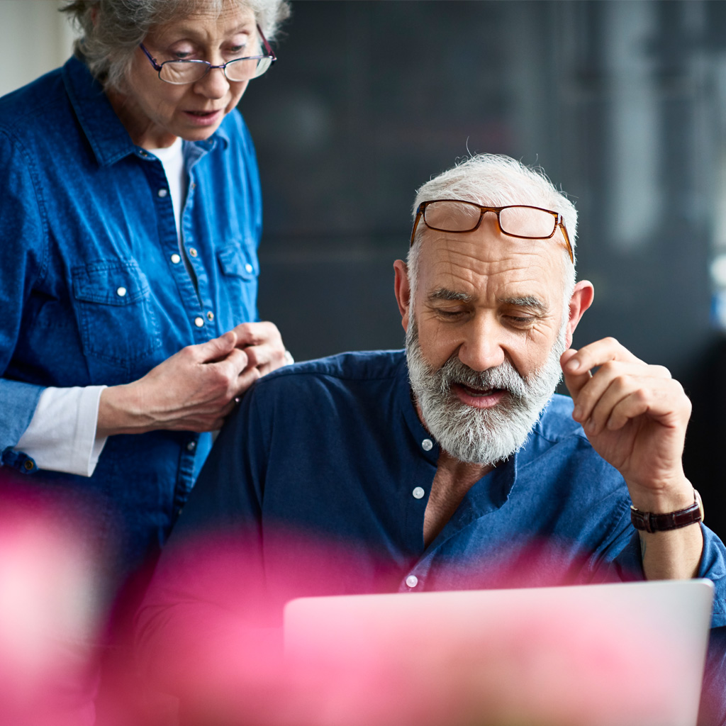 Two older people with grey hair, wearing blue denim shirts looking at a silver latop. The older man has his galsses on his head and is sitting down, the female is standing up and wearing glasses on the bridge of her nose,. Two older people with grey hair, wearing blue denim shirts looking at a silver latop. The older man has his galsses on his head and is sitting down, the female is standing up and wearing glasses on the bridge of her nose,.