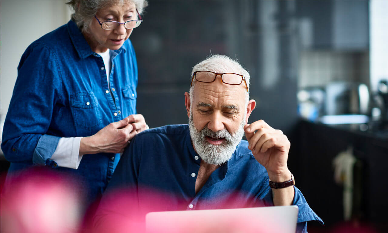 Two older people with grey hair, wearing blue denim shirts looking at a silver latop. The older man has his galsses on his head and is sitting down, the female is standing up and wearing glasses on the bridge of her nose,.