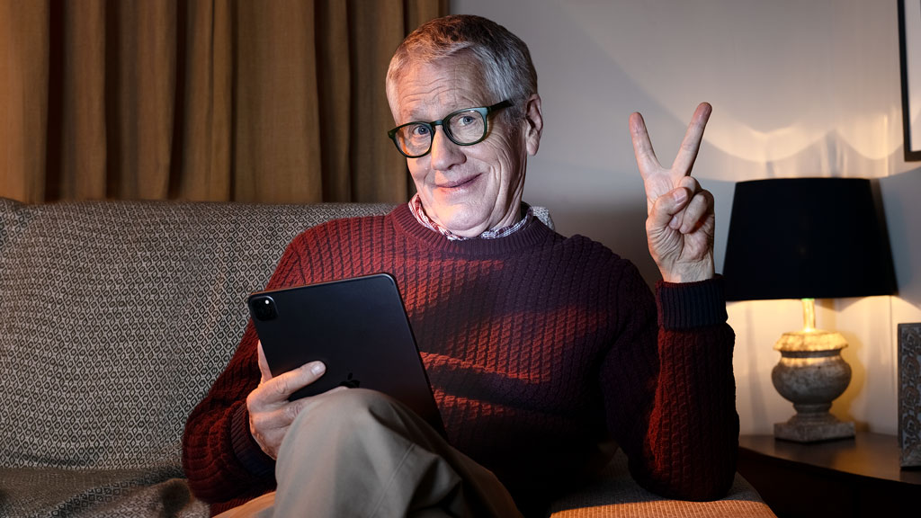 Older man sitting on a sofa with a tablet making the 'peace' sign Older man sitting on a sofa with a tablet making the 'peace' sign