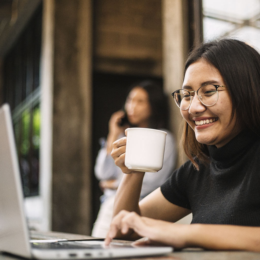 A women with brown hair, wearing glasses, sitting at a table and smiling at a laptop, with a mug in her hand A women with brown hair, wearing glasses, sitting at a table and smiling at a laptop, with a mug in her hand