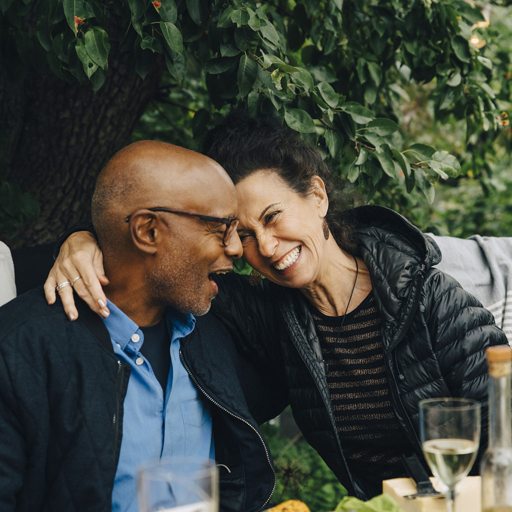 A women in a black coat, with her arm around a man wearing black rimmed glasses, sat in the garden smiling at each other A women in a black coat, with her arm around a man wearing black rimmed glasses, sat in the garden smiling at each other