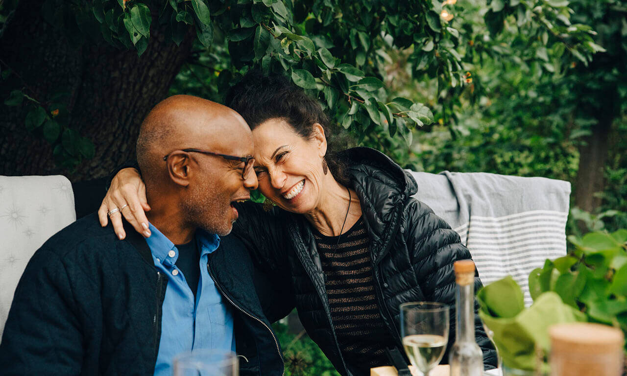 A women in a black coat, with her arm around a man wearing black rimmed glasses, sat in the garden smiling at each other